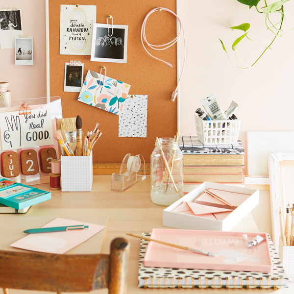 Secretária coberta com revistas, canetas, marcadores e papéis. Na parede em frente à mesa há um quadro de avisos coberto de anotações e fotos.