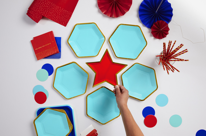 Uma mão com um prato em forma de estrela vermelha no centro de um prato semelhante a um patriota, um prato hexagonal azul claro e dourado, ventiladores de papel azul vermelho e escuro.