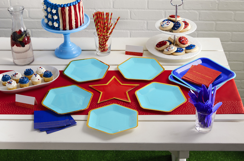 Uma mesa de festa patriótica com placas hexagonais azuis e douradas, pratos em forma de estrela vermelha, ventiladores de papel azul vermelho e escuro.