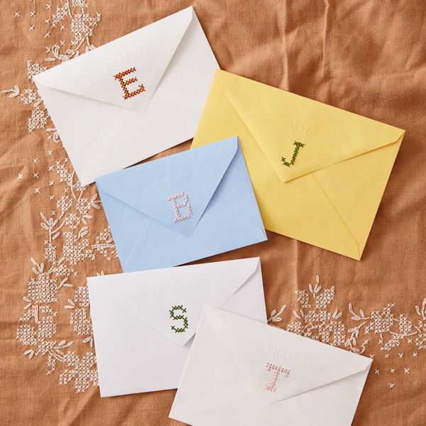 Os cinco envelopes de cartões são costurados nas abas traseiras na parte traseira de cad a-e m-monograma.< pan> itens de artesanato, como flor de papel, barbante, tinta e tesoura, estão espalhados na bancada de Woodgrain.