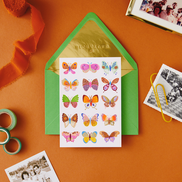 Um cartão com três fileiras de borboletas de diferentes cores e desenhos na frente. Abaixo do cartão há um envelope verde brilhante com folha de ouro.