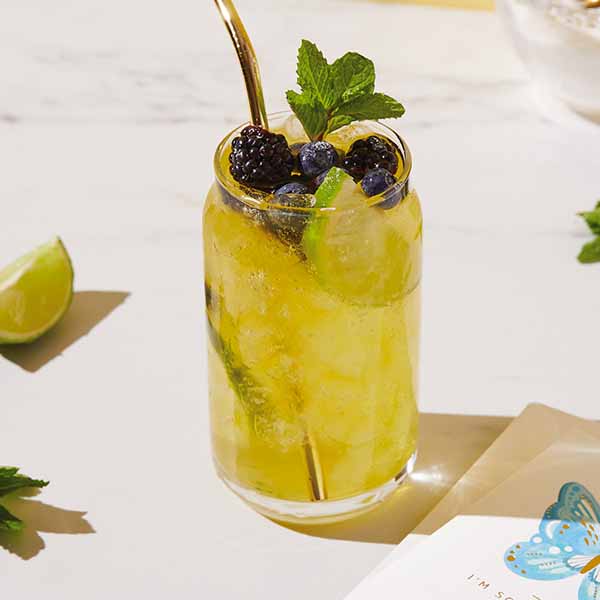 Um copo de refrigerante de chá verde com amora, limão e hortelã, uma palha de metal de placa dourada é presa e é colocada em um balcão de mármore branco e cinza.