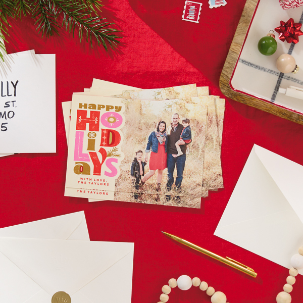Sobre uma mesa coberta por uma toalha vermelha brilhante está uma pilha de cartões fotográficos exclusivos e festivos, cercados por envelopes, canetas e folhagens festivas.