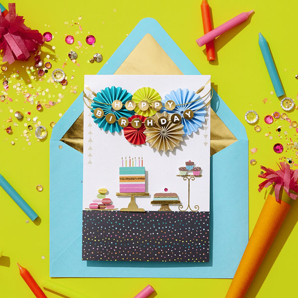 2278_II_Um cartão de aniversário de uma amiga decorado com bolo e guirlanda em envelope azul, com confetes e serpentinas ao fundo.
