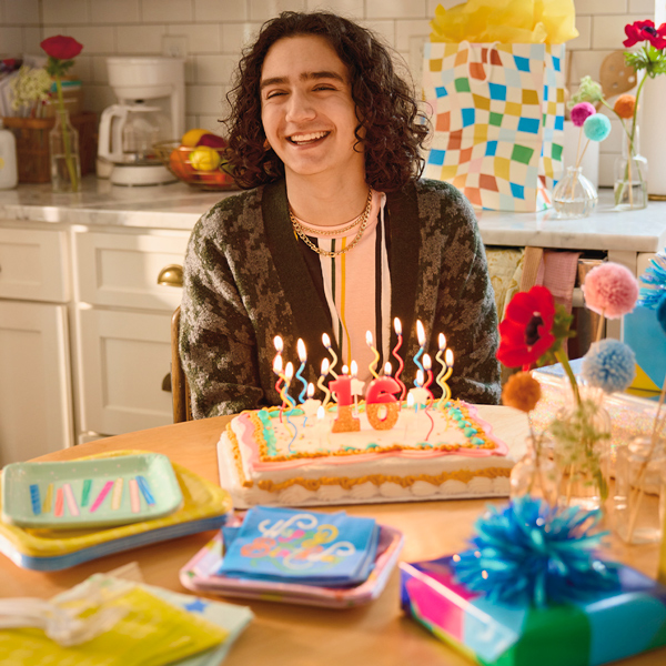 Atrás do bolo de aniversário fosco, um adolescente fica sentado em uma mesa de cozinha.