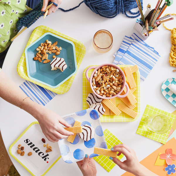 Uma pequena foto de uma coordenação de mesa brilhante e colorida para uma festa de artesanato. Uma mulher estende a mão para oferecer um prato de doces a outra mulher que está tricotando do outro lado da mesa.