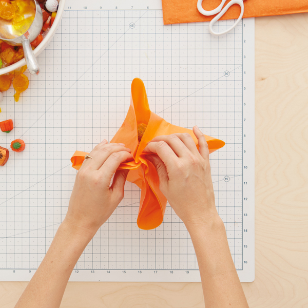 Halloween-Pumpkin-Treat-Bag-3: uma mão que dobra um canto de papel laranja quadrado no meio, como se cercado por montanhas doces.