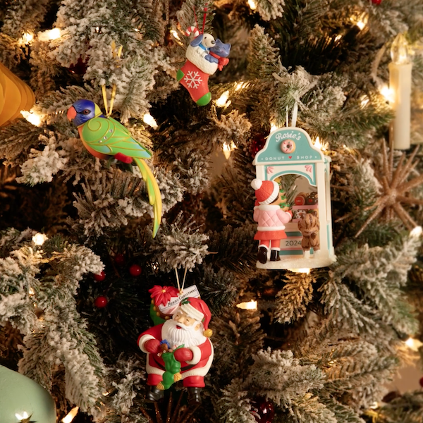 Quatro enfeites comemorativos exclusivos, disponíveis apenas para membros do Keepsake Ornament Club em 2024, estão pendurados na árvore de Natal iluminados por luzes brancas e velas em estilo vintage.