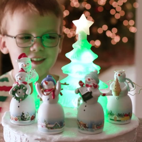 Um menino de pijama e óculos listrados de vermelho, verde e branco sorri para um enfeite de mesa representando uma árvore de Natal cintilante e cinco bonecos de neve.
