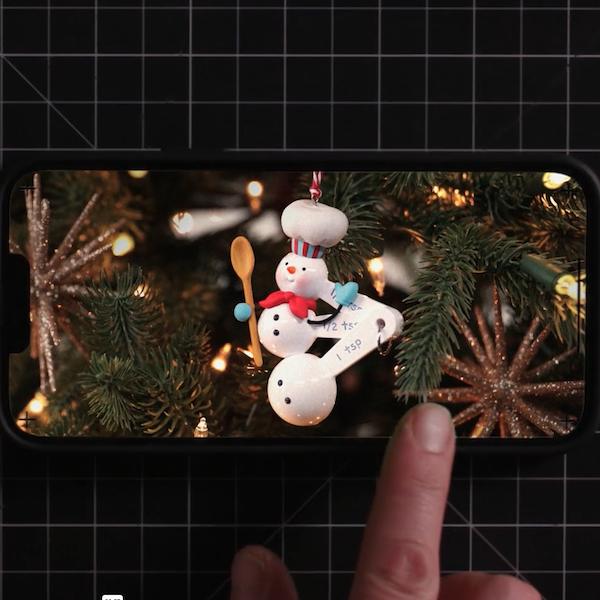Um smartphone é colocado em um tapete de corte com uma grade branca e preta, com uma colher de pau, e uma foto de um ornamento comemorativo do boneco de neve usando um chef é exibido no smartphone.