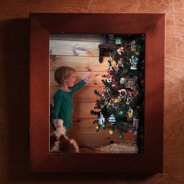 Uma foto emoldurada de um menino surpreso de 3 a 4 anos, vestindo pijama verde, segurando seu bichinho de pelúcia favorito em uma mão e segurando uma árvore de Natal totalmente iluminada e decorada na outra. .