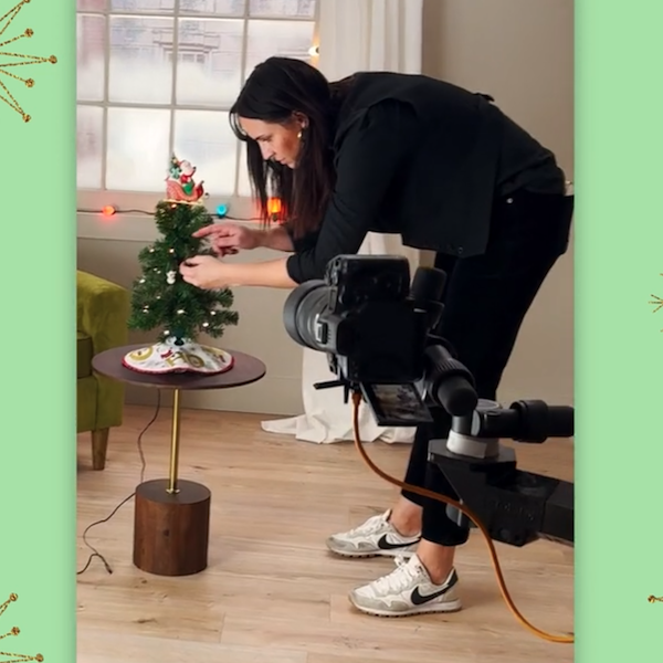 Um fotógrafo vestido de preto se inclina para ajustar os enfeites pendurados em uma pequena árvore de Natal. Em primeiro plano, uma câmera digital profissional está acoplada a uma câmera Foley, indicando que a cena está sendo filmada.