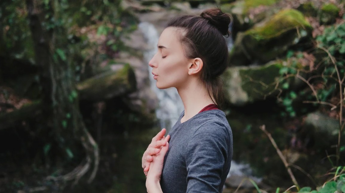 Uma mulher fica em frente a uma cachoeira, fecha os olhos e faz exercícios respiratórios.